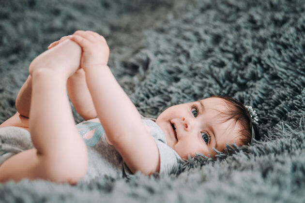 脚趾可爱的蓝眼睛婴儿躺在卧室的灰色床单上 抱着她的腿护理快乐可爱