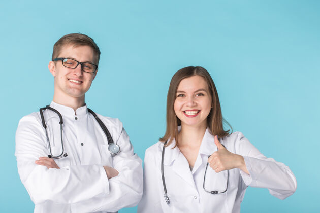 从业者两位最聪明的专业微笑医生穿着白大褂 竖起大拇指站在蓝色的表面上女性医院男性