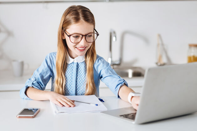 自我技术使用智能在阳光明媚的厨房里 一个专注的小学生坐在桌子旁写着她的文章 在网上寻找一些提示童年科技手机