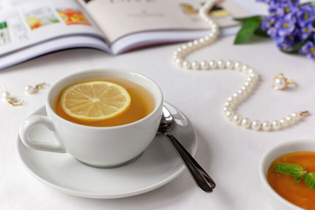 书白色床单上放着一杯绿茶 柠檬 蜂蜜和薄荷 床单上有插图杂志 珍珠项链 耳环和戒指茶杯子柠檬