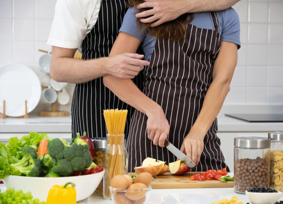 围裙夫妻俩在厨房准备健康食品雌性在砧板上切苹果 雄性拥抱并站在她身后夫妇健康美食和家常菜一起概念碗砧板胡萝卜