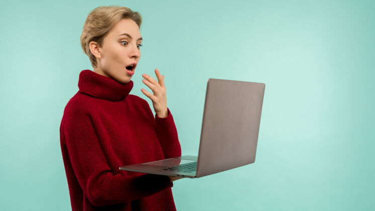 女性一个穿着毛衣的快乐金发女人在蓝色背景下张嘴看笔记本电脑屏幕-图片休闲学生手指