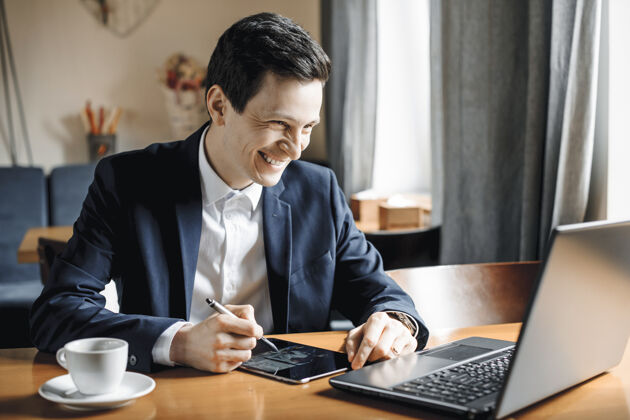 工作场所一个英俊的商人坐在桌子上用笔记本电脑工作 一个平板电脑微笑着看着笔记本电脑的屏幕设计师电脑休闲