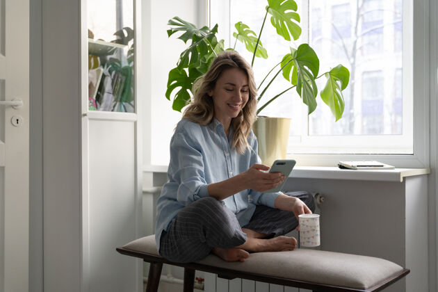 女人微笑的女人在家里端着一杯咖啡 用手机智能手机 在社交网络上聊天智能怪物文本