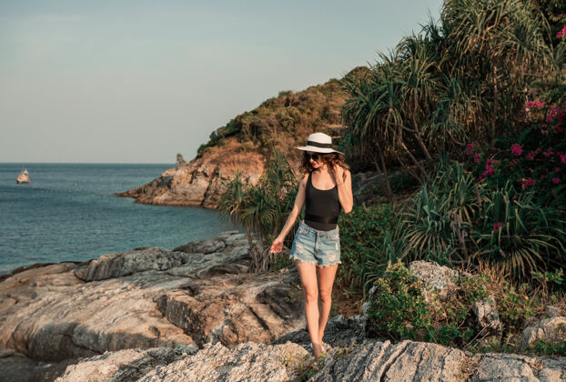 比基尼在热带海滩的一块大石头上穿着柳条的梦幻女人帽子概念暑假旅行的乐趣女人年轻阳光