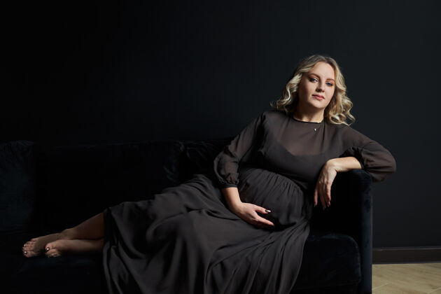 时尚孕妇在一个优雅的黑色礼服摆姿势室内工作室黑墙背景腹部期望妈妈