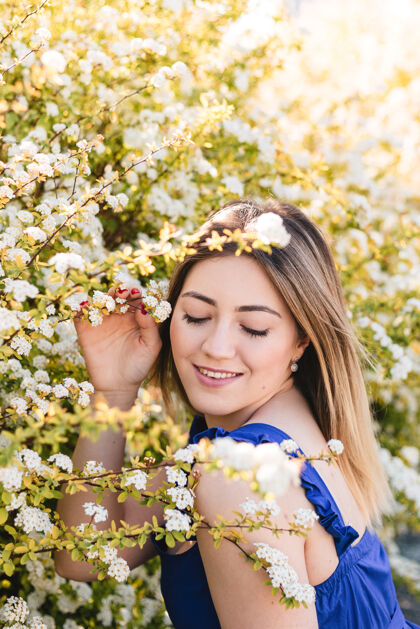 花美丽的时髦女人在盛开的灌木中的感性平静的肖像 白色的花朵绣线菊空间时尚花园里穿着蓝色裙子的金发女人春天肖像女人