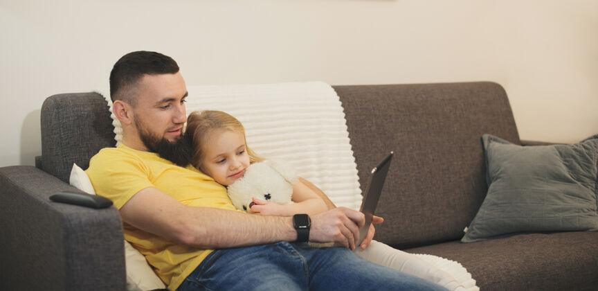 父母可爱的女孩躺在她父亲的肩膀上 拥抱着一只泰迪熊 同时看着平板电脑一起男人谎言