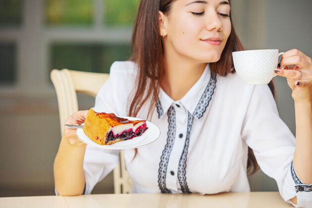 独自年轻漂亮的快乐女孩正在吃早餐咖啡和芝士蛋糕坐在桌边女士蛋糕大笑