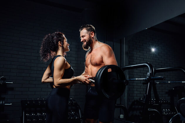 运动侧视图英俊的胡须男子和美丽的女子微笑着看着对方 而与杠铃在黑暗的健身房锻炼运动员努力男人