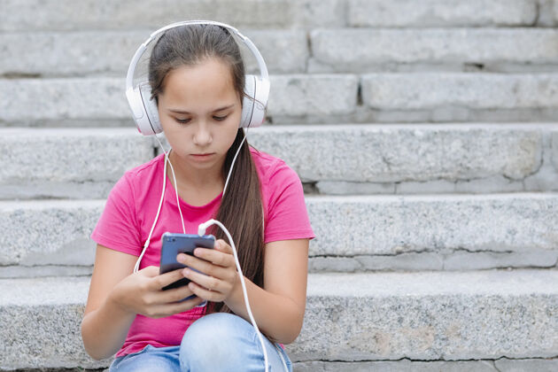 人戴着耳机的女孩坐在台阶上 手里拿着一部智能手机手长头发 粉色t恤 蓝色牛仔裤女性手机步骤
