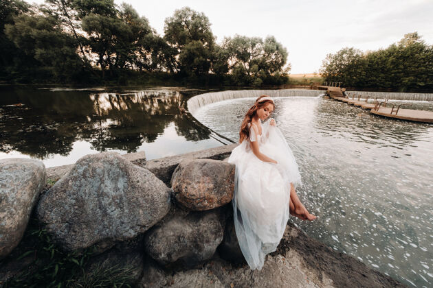 女孩一位穿着白色礼服 戴着手套 赤脚的优雅新娘正坐在公园的瀑布旁 享受着大自然河流山河户外