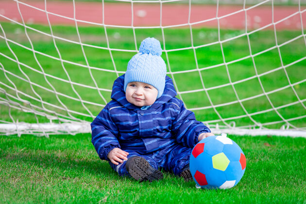 户外穿着蓝色工作服的婴儿坐在球门前的足球场上可爱球员婴儿