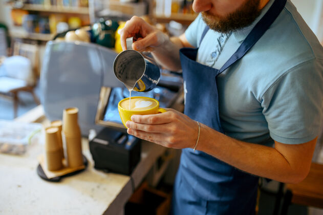 准备围裙里的男咖啡师往咖啡里倒泡沫咖啡馆男在自助餐厅做新鲜的浓缩咖啡 酒吧柜台的服务员室内机器男人