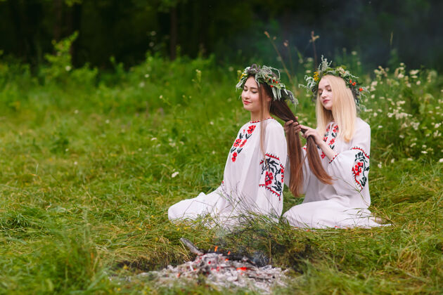 传统仲夏二穿着斯拉夫衣服的女孩在火炉旁的头发上编辫子斯拉夫女孩田野