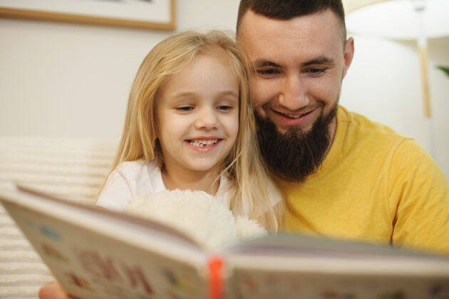爸爸一位可爱的留着胡子的年轻父亲的特写照片 早上母亲在家工作时 他和女儿在看书父母胡子金发