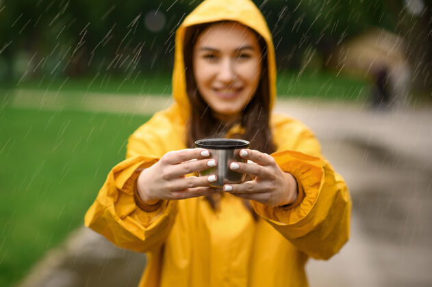 雨穿着雨衣的女人端着一杯热茶 在雨天的夏日公园里 透过窗外的景色一个人女人穿着雨衣走在小路上 雨天在巷子里自由季节气候
