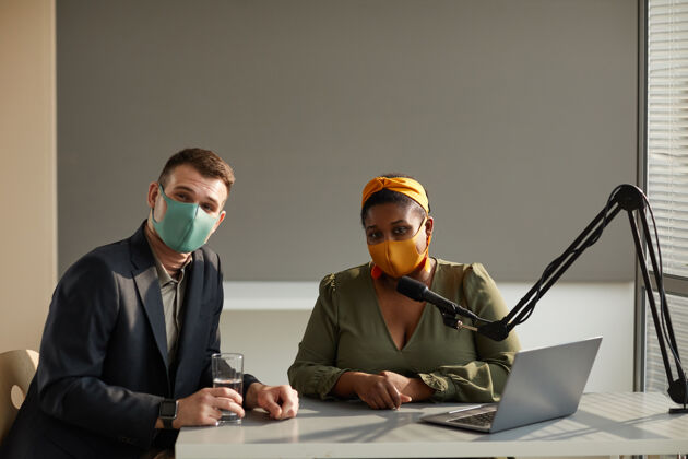 商务人员年轻的电台dj和嘉宾戴着防护面具坐在桌子旁 在电台采访时看着前方的画像计算机使用笔记本电脑伙伴关系