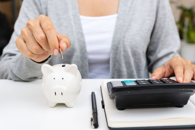 税务特写镜头中 一位女士一边用计算器 一边把硬币放在小猪存钱罐里主页.家庭费用和节省概念昂贵女性支票