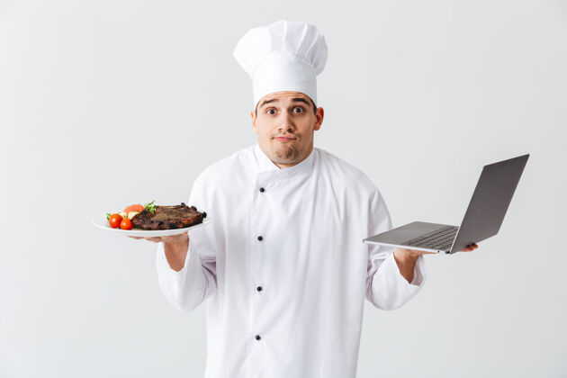 菜肴困惑的厨师穿着厨师制服站在白色的墙上 拿着笔记本电脑 展示着一道菜人男性互联网
