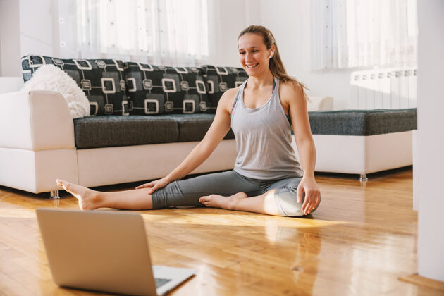 健美操面带微笑的肌肉健身教练坐在家里的地板上 做伸展运动锻炼她正在笔记本电脑上进行在线课程强壮健康的生活方式女运动员