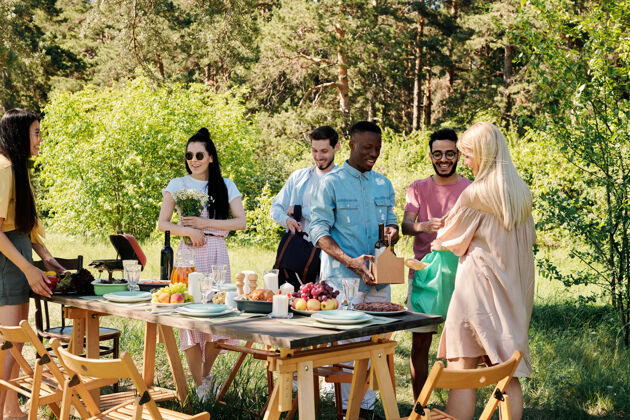 生活方式一群年轻欢快的国际友人穿着休闲服站在松树下的桌子旁 放上食物 饮料和鲜花友谊休闲服户外