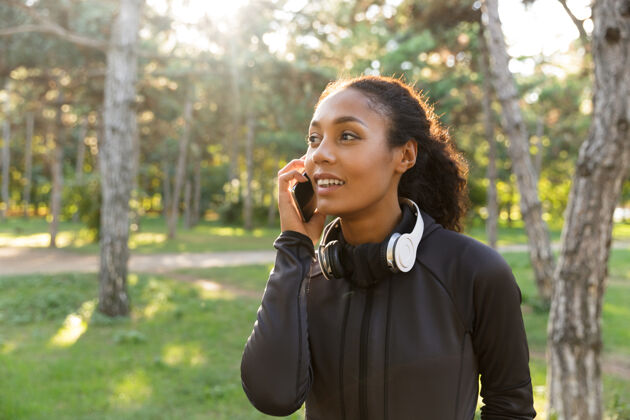锻炼一个20多岁的快乐女人穿着黑色运动服 戴着耳机 一边在绿色公园散步一边用手机跑步女孩耳机