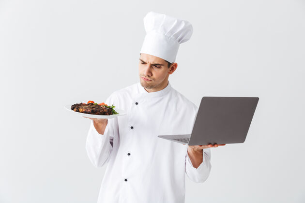 厨师正经的厨师穿着制服站在白色的墙上 拿着笔记本电脑 展示着一道菜盘子年轻人沟通