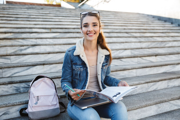 户外微笑的年轻女子穿着夹克坐在室外的楼梯上 做作业 学习年轻休闲白种人