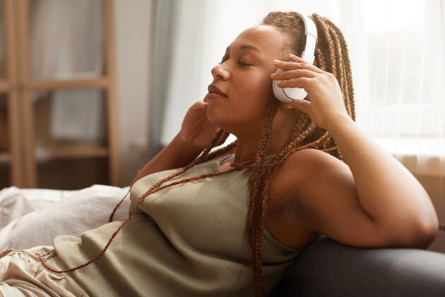 卧室非洲年轻女子戴着无线耳机坐在家里的沙发上欣赏音乐家庭室内放松耳机