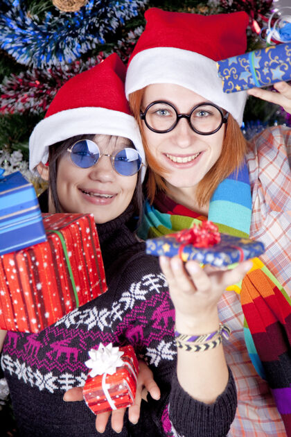 年轻人两个漂亮的女孩在圣诞树旁戴着圣诞帽 手里拿着礼物盒子无忧无虑红发