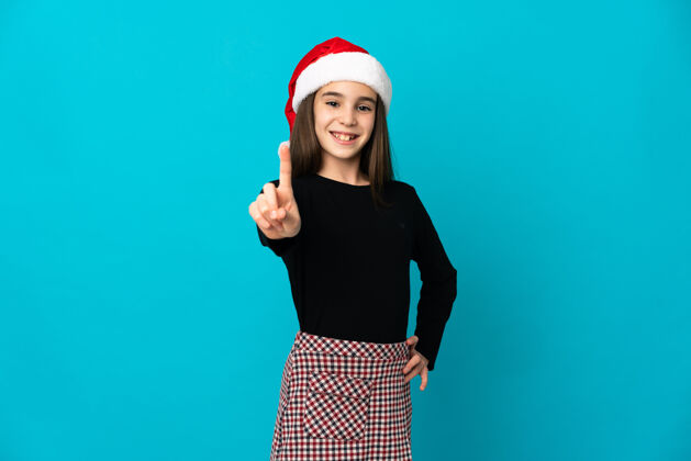 1戴着圣诞帽的小女孩在蓝色背景上孤立地展示和举起一个手指111