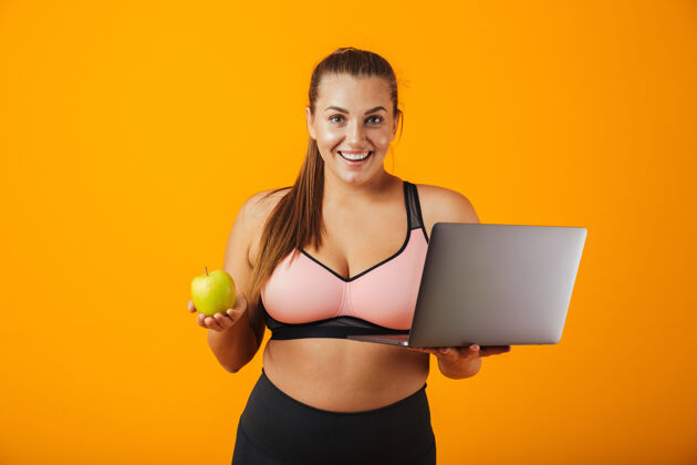 肥胖一个穿着运动服的快乐超重的年轻女子的肖像 孤立地站在黄色的墙上 用笔记本电脑 拿着绿色的苹果放松笔记本电脑运动