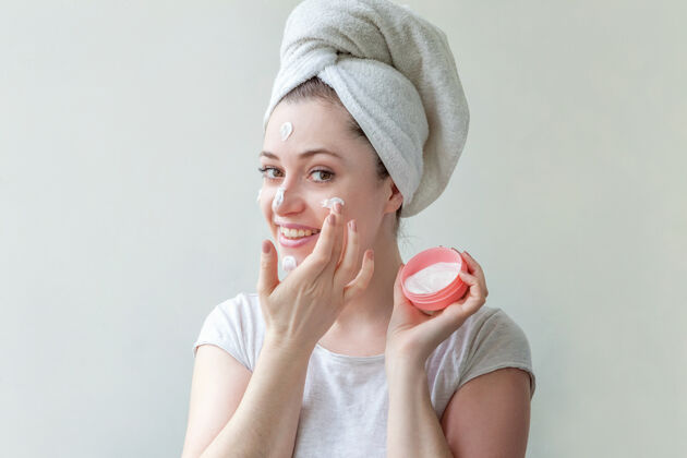 皮肤科在头像上用毛巾裹着的小美女在脸上涂上白色滋养面膜或乳霜 隔离在白色背景上美容美容师皮肤