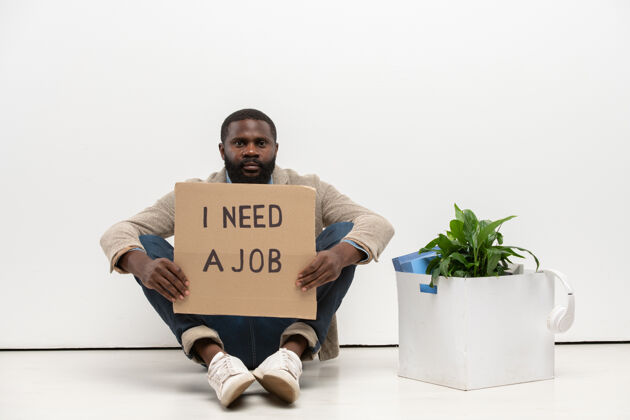 信息非洲裔的年轻失业者拿着一张纸板说他需要一份工作 坐在办公室白墙边的地板上男子举行求职