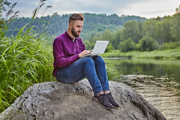 户外年轻的大胡子男人坐在河边的石头上 膝上放着笔记本电脑 他看着屏幕 在键盘上打字人自由职业者人