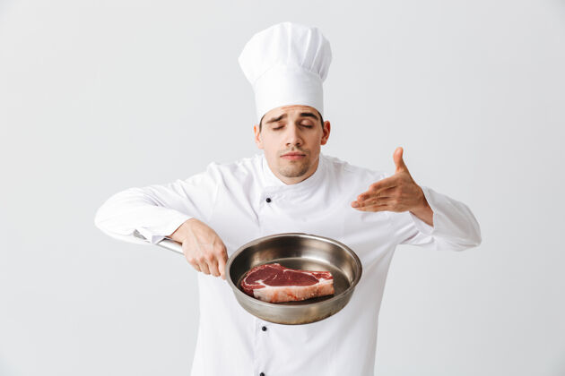 食谱开朗的厨师身着制服 在白墙上展示一个生牛排的煎锅晚餐烧烤专业