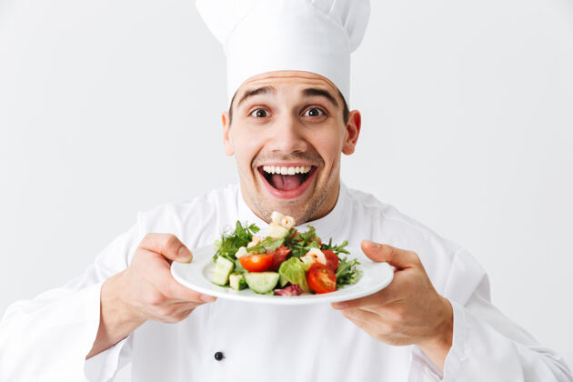 菜肴兴奋的男厨师穿着制服 把新鲜的蔬菜沙拉放在隔离在白色墙上的盘子上信心盘子烹饪