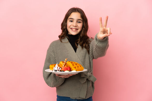 可爱小女孩拿着粉红色背景上孤立的华夫饼 微笑着展示胜利的标志女孩甜点和平