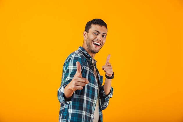 手势一个穿着格子衬衫的快乐的年轻人孤立地站在橙色的墙上 指着摄像机思考表演方格