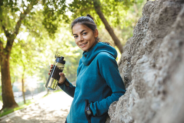 肖像特写镜头：一位穿着运动服 手持一瓶水在户外锻炼的迷人的年轻健身女士饮酒夏天女人