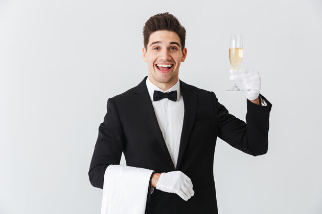 携带身着燕尾服的微笑年轻侍者的画像在白墙上为您奉上一杯香槟微笑酒精工作人员