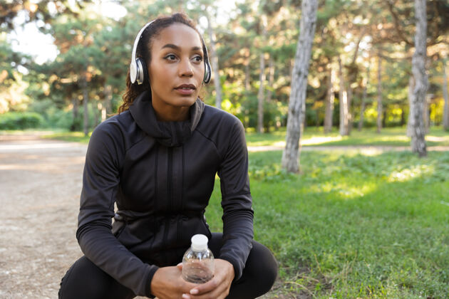 运动一个20多岁的美国年轻女子穿着黑色运动服 戴着耳机 拿着水瓶在绿色公园里散步无线美国人户外
