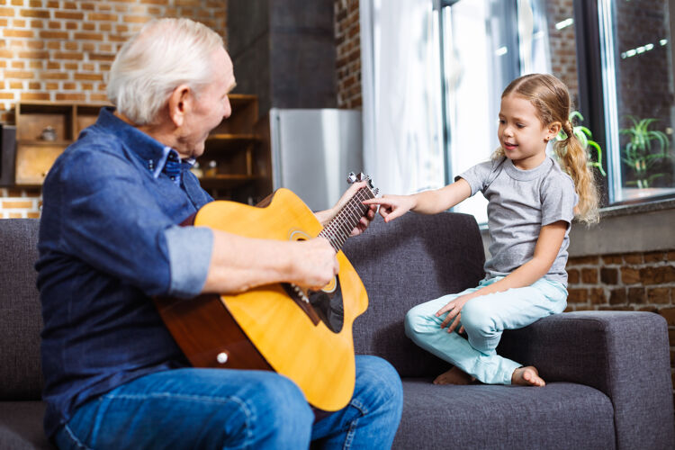 男性快乐的老人在家为他微笑的小孙女弹吉他老人快乐沙发