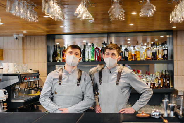 员工在流感大流行期间 两个戴着口罩和制服的时髦调酒师站在吧台后面在流感大流行期间 餐厅和咖啡馆的工作口罩服务防护