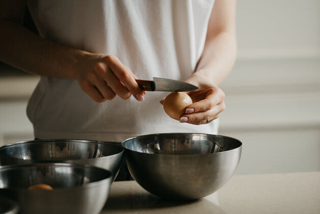面包房厨房不锈钢碗上方 一名年轻女子用刀打碎蛋壳的照片烹饪准备鸡蛋
