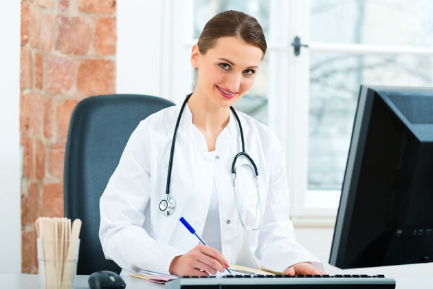 电脑年轻的女医生坐在诊所窗户前的书桌旁写文件或档案医生职业听诊器