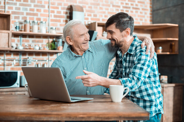 房子开朗善良的老人微笑着拥抱他的儿子 同时学习如何使用笔记本电脑进步老年人老年人