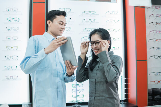 屏幕一位女顾客试着戴上眼镜看平板电脑屏幕 而一位店员拿着平板电脑站在眼镜架展示柜的背景下戴眼镜商顾客