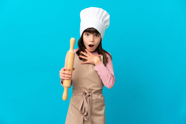厨师小女孩拿着擀面杖孤零零地站在蓝色的墙上 看着右边 既惊讶又震惊卷女孩员工